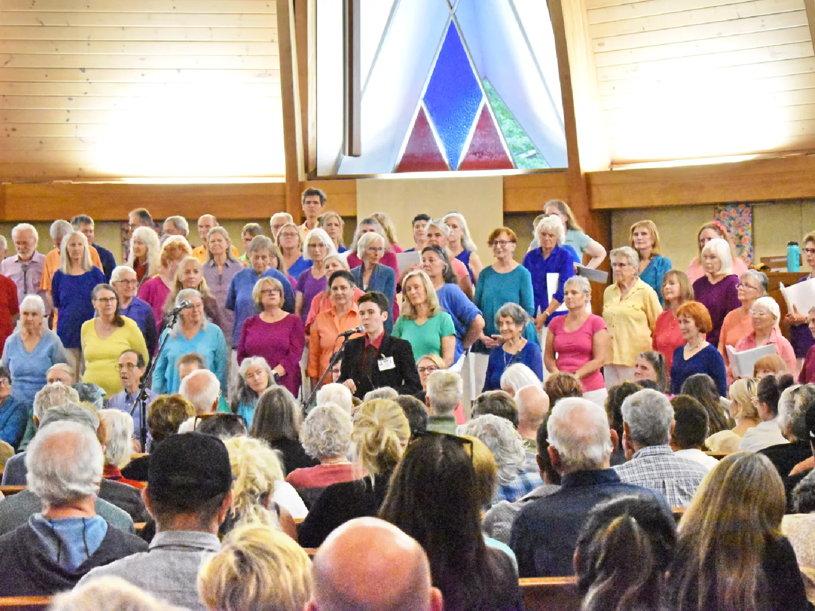 A volunteer is speaking at a church in front of a large group of people with a large number of people in colorful shirts standing behind the volunteer.