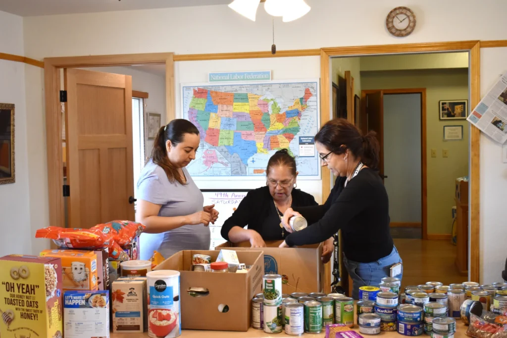 Three women fill a box of non-perishable food items with a map of the United States behind them