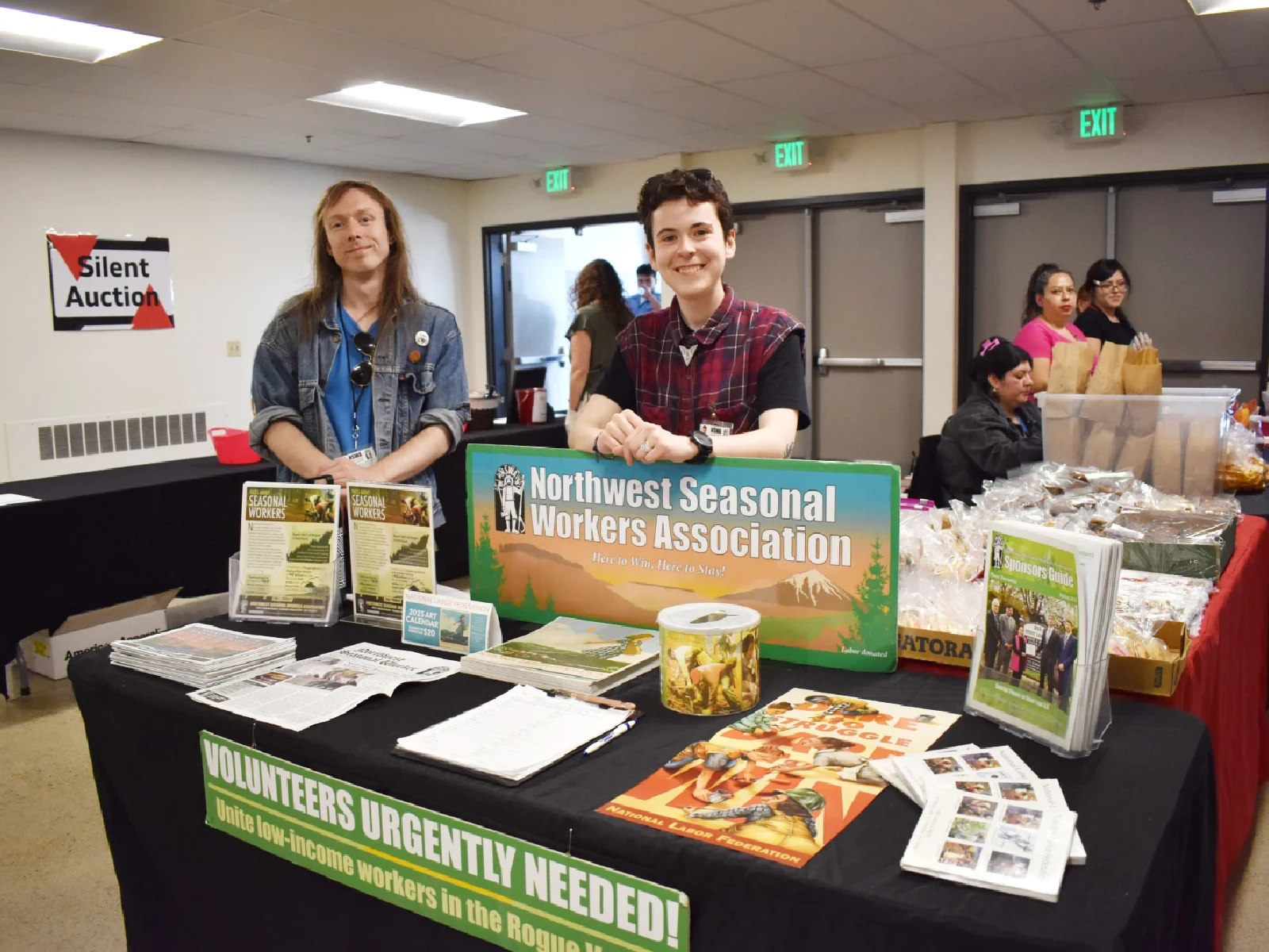 Two volunteers stand behind a table with information displayed about the organization. In the background there are other people staffing tables with food.