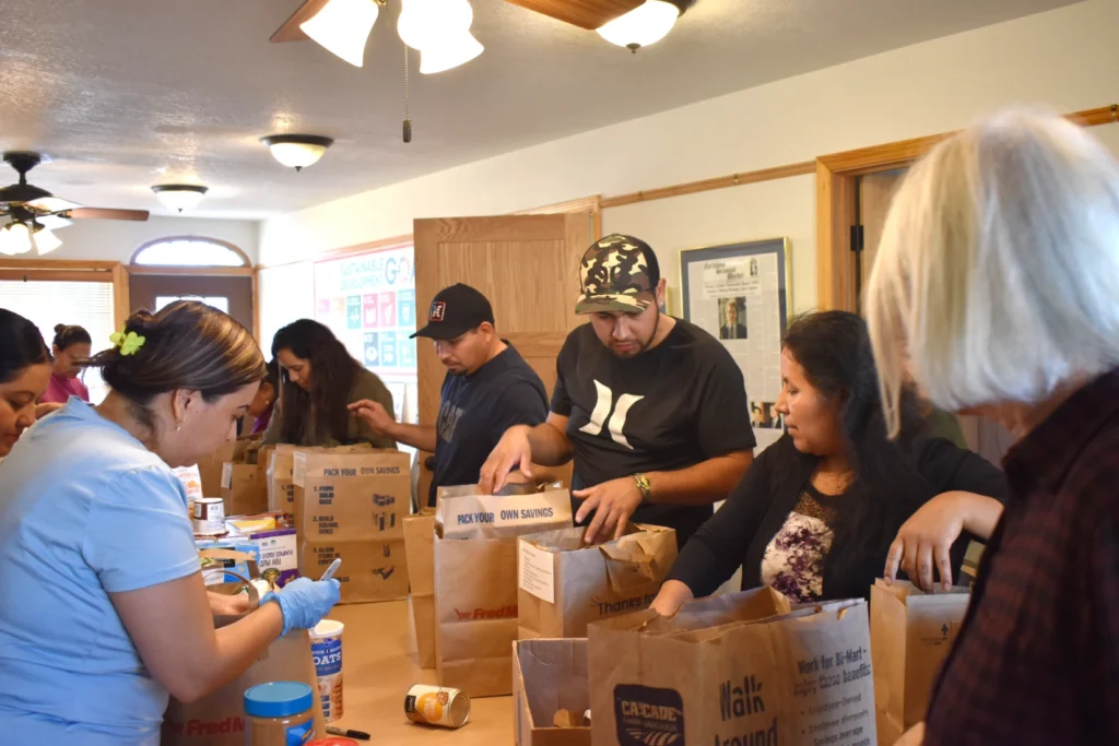 A group of people stand around a table in an office and fill paper bags with canned food items