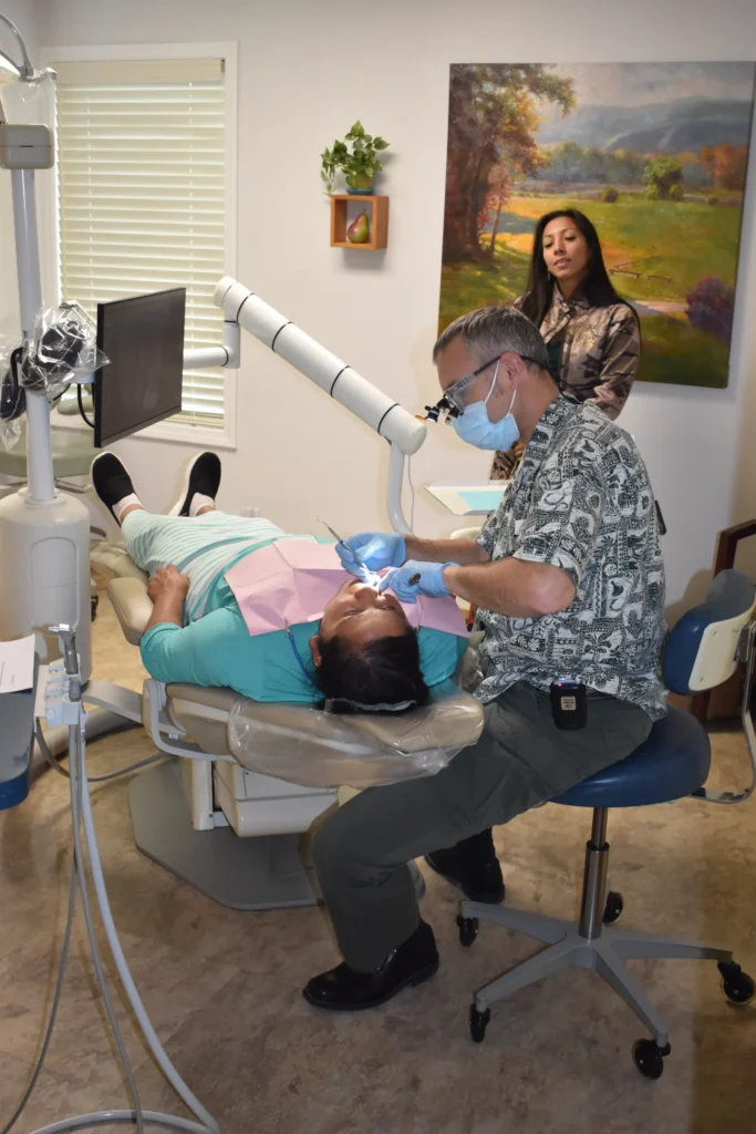 A dentist does an exam on a patient while another volunteer stands in front of a painting in the room.