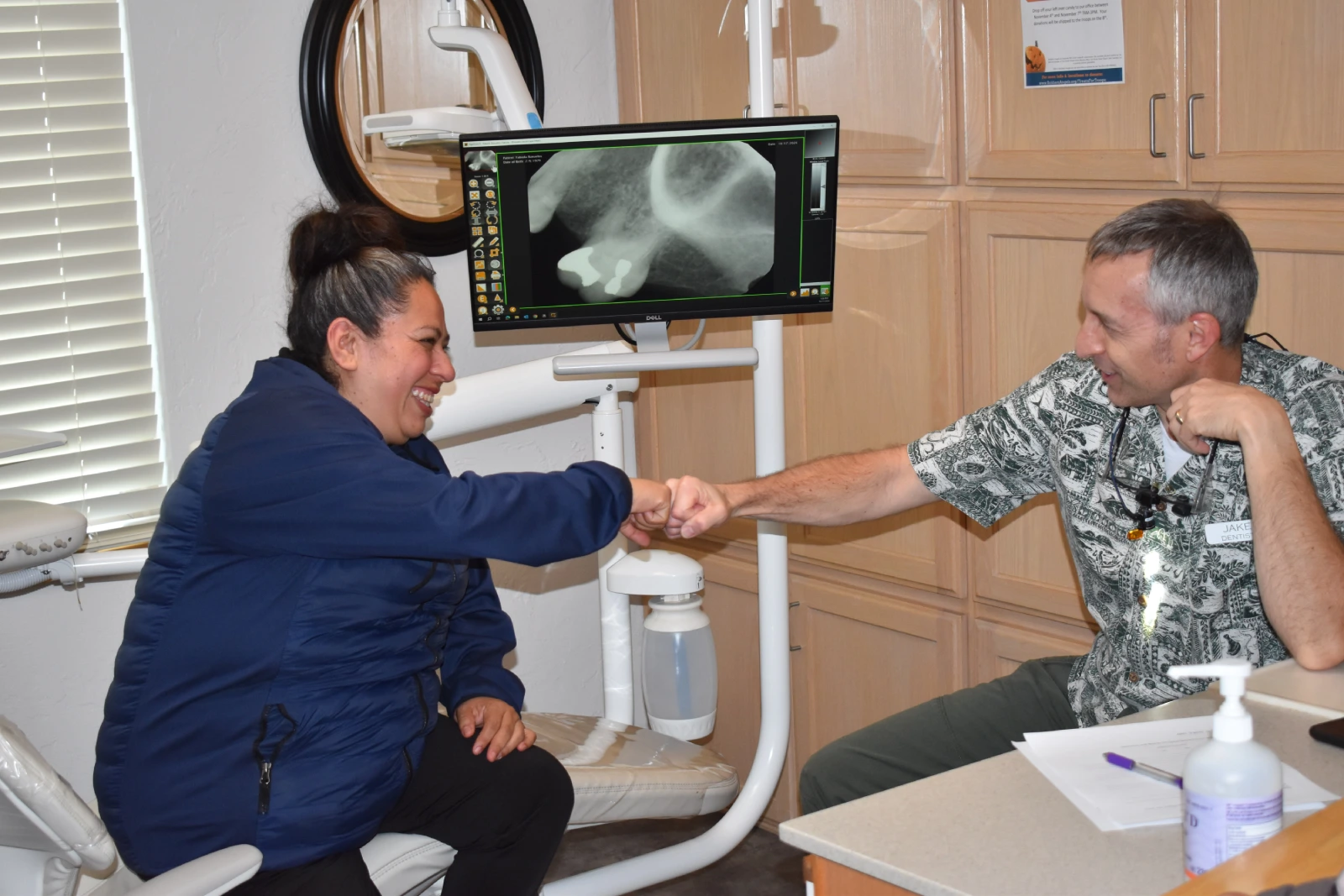A woman sitting in a dental office does a fist bump with a dentist and both are smiling.