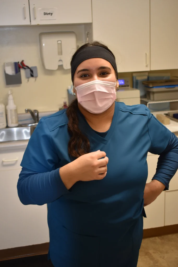 A dental assistant who is wearing a mask stands in a dental office