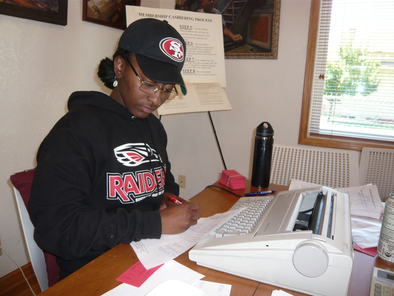 A college student wearing a baseball hat writes on a sheet of paper as she sits in front of a typewriter in an office