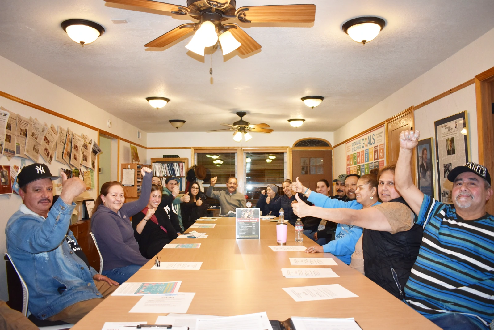 A group of members are sitting around a table in an office and they are all giving the thumbs up