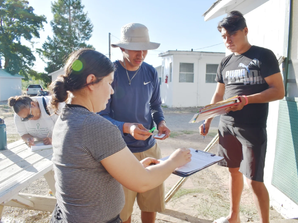 A young woman and young man with clipboards speak with a farm worker at a farm labor camp.